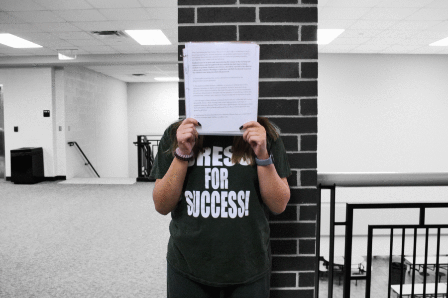 An anonymous student stands outside the office wearing Eagle Greens, holding up the Code of Conduct with dress code rules.Eagle Greens are used as the alternative to a inappropriate outfit and a punishment to those not following the dress code rules. 