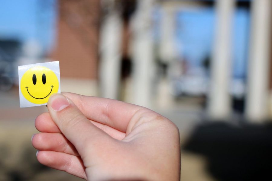 Basking in the sunshine, senior Emma Hutchinson holds up a smiley face sticker she received from a Hope Squad member. The Hope Squad handed out stickers and candy while greeting students and teachers entering the high school the morning of Feb. 1. This week is Hope Week, which includes creative dress up days and involvement for all students.