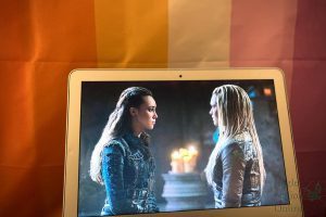 In front of a lesbian flag, the CWs The 100 plays on a MacBook. Lexa and Clarke had a romantic relationship in the second and third seasons of the show. Lexa died taking a bullet meant for Clarke after they made their relationship official in season 3 episode 7, a prime example of the Bury Your Gays trope.