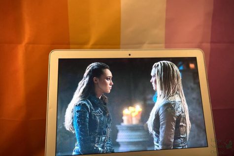 In front of a lesbian flag, the CWs The 100 plays on a MacBook. Lexa and Clarke had a romantic relationship in the second and third seasons of the show. Lexa died taking a bullet meant for Clarke after they made their relationship official in season 3 episode 7, a prime example of the Bury Your Gays trope.