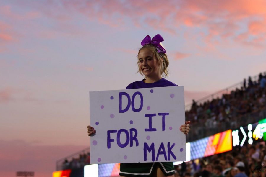 Performing a stunt, junior Kendall Smith holds a sign in honor of fellow cheerleader Makayla Noble. Noble recently injured her spinal cord in a tumbling accident and is currently hospitalized. Community members, staff and students wore purple to the game Sept. 24 in her honor.