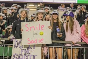 Holding up a 'Smile for Sadie' sign, senior Gavin Teague and junior Alyssa Milner stand in the Talonette section. The Talonettes performed a dance dedicated to their teammate Sadie Neddo. A prayer was held at the end of the game to honor her. 